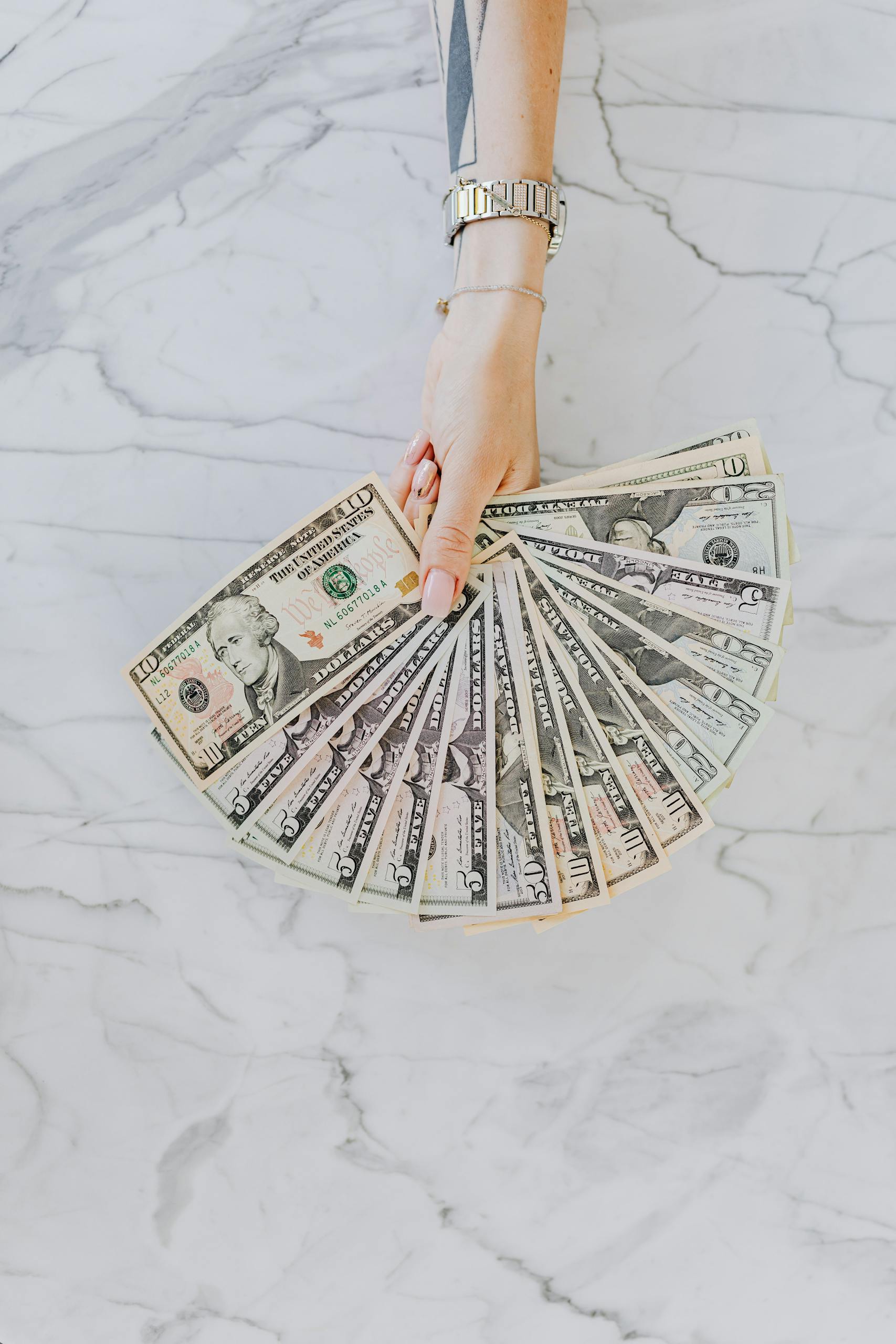A hand with wristwatch holding a fan of US dollar bills on a marble surface, symbolizing wealth and financial abundance.